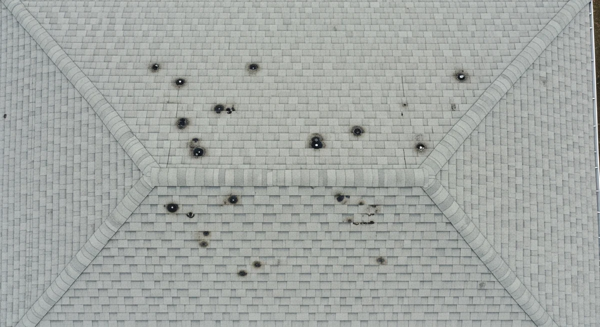 Aerial view straight down on a pale grey hip roof, with roughly twenty small dark circular hail bruises scattered sparsely and unevenly across two slopes and large areas of shingle left untouched.