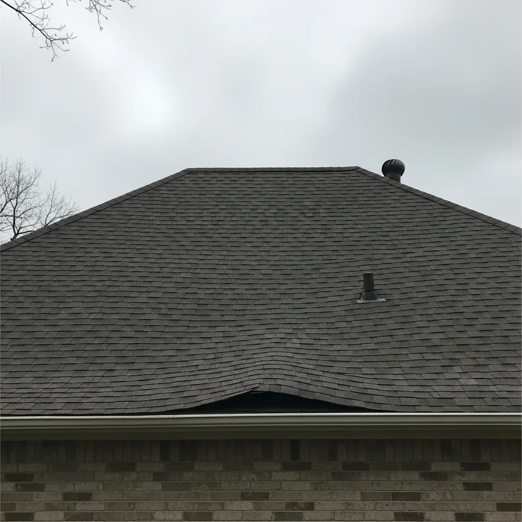 Ground-level view of a hip roof with a visibly sagging, wavy dip in the shingle courses near the eave, on an overcast day, with a roof vent stack and bare tree branches in frame.